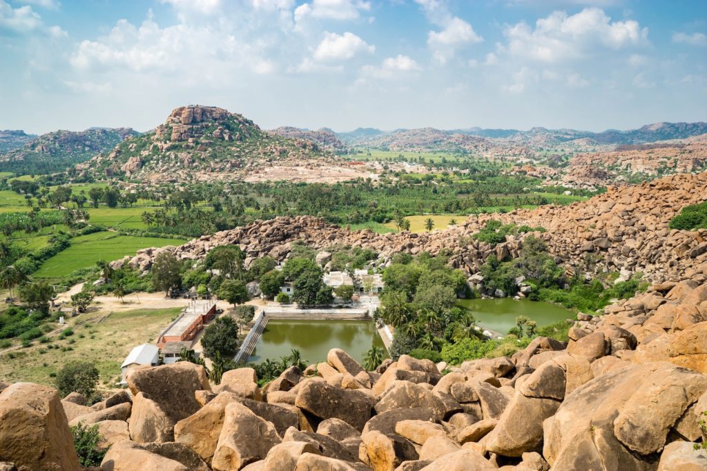 A scenic landscape featuring large boulders in the foreground, a serene body of water, clusters of buildings, lush greenery, and distant hills under a cloudy sky.