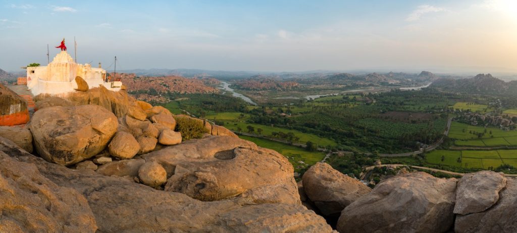 A panoramic landscape showcasing a temple with a red flag atop a rocky outcrop, overlooking sprawling green fields and distant mountains at dusk.