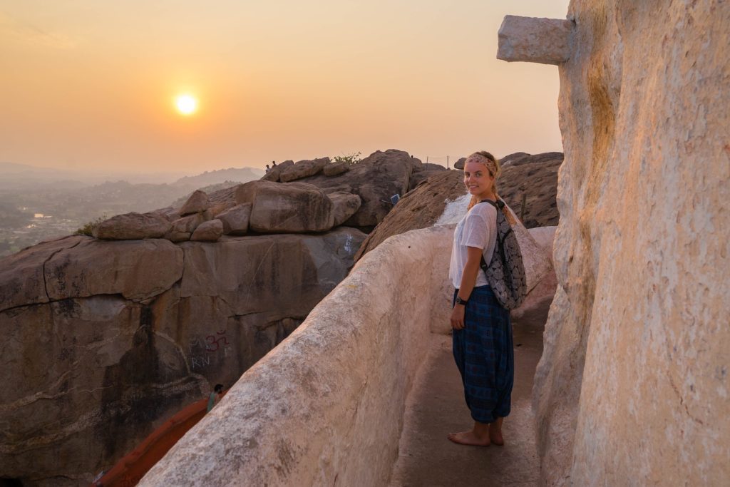 A person stands on a rocky path with a sunset in the background. They wear casual clothing, smile, and appear relaxed in a scenic landscape.