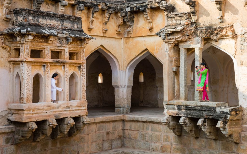Two people are standing on separate balconies of an ancient, ornately carved stone structure. The person on the right is wearing colorful traditional attire.