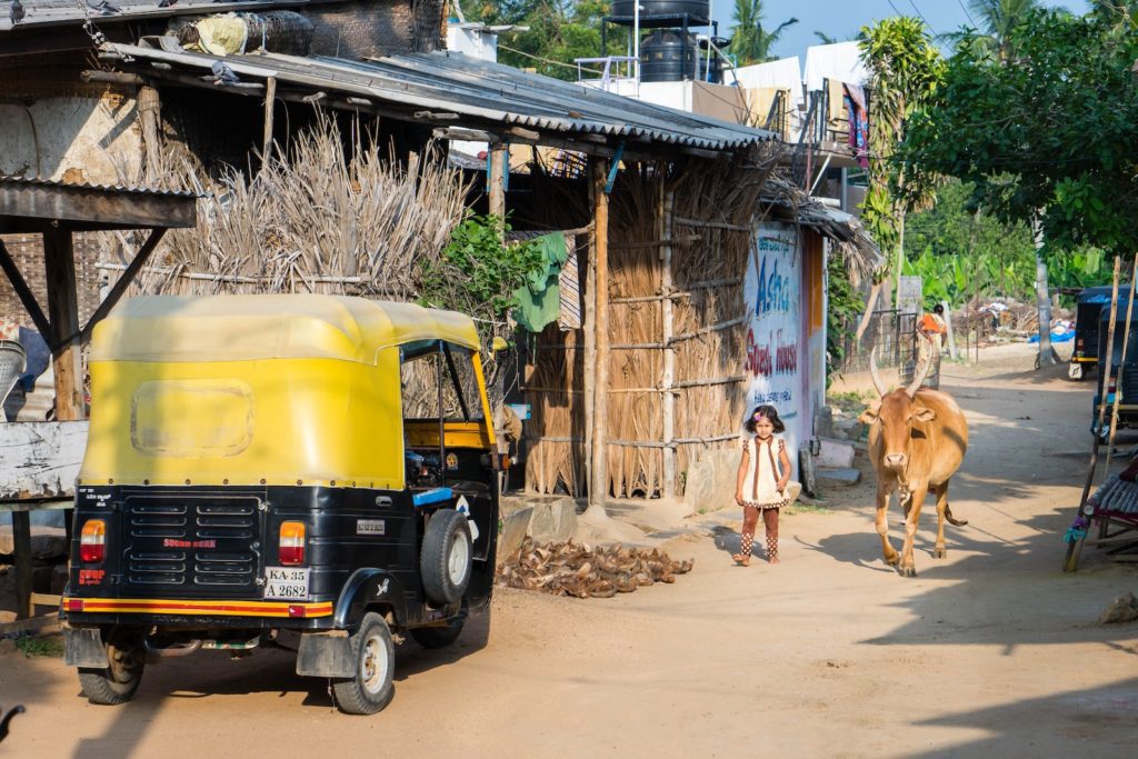 A rural street scene with a yellow auto-rickshaw parked, a cow walking, and a child standing in the middle. Thatched huts and trees are visible.