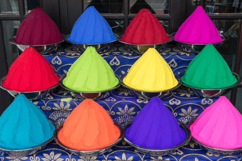 This image shows twelve vibrant, conical piles of powdered color on trays, arranged on a blue patterned cloth, likely for a celebration like Holi.