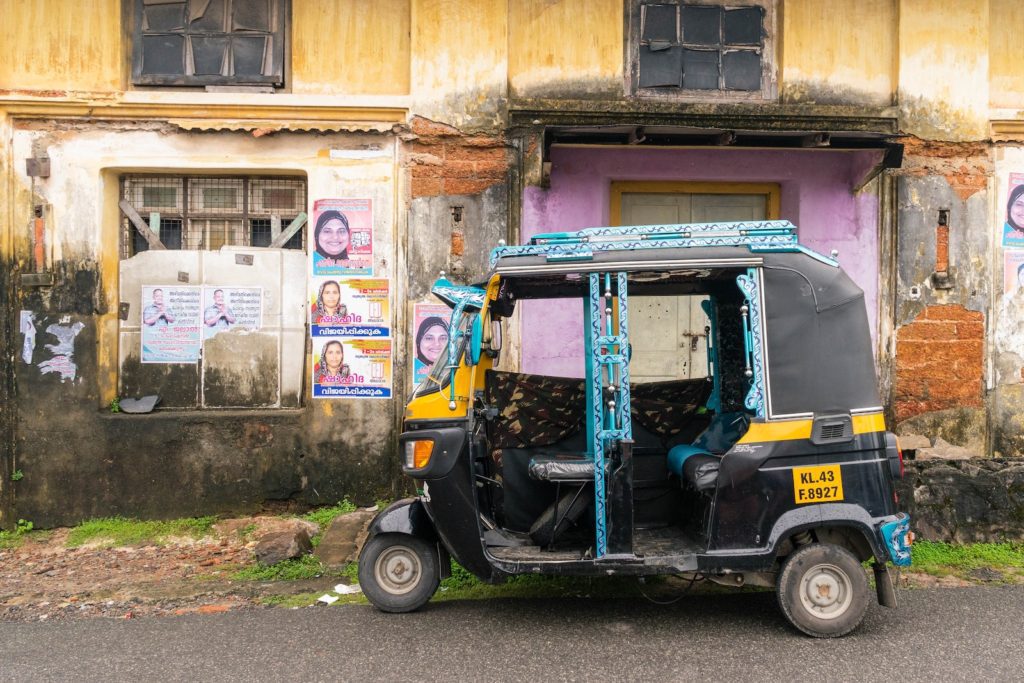 An auto-rickshaw parked in front of a weathered building with yellow walls. Election posters with faces of candidates are visible on the wall.