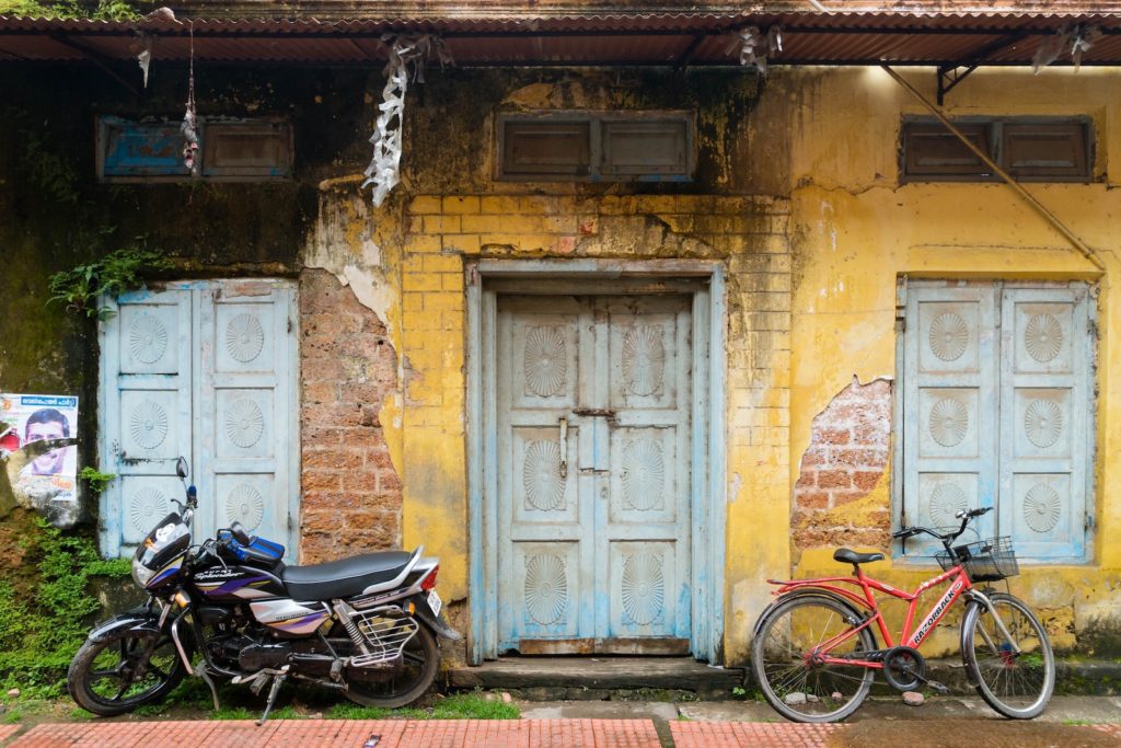 An old building with weathered blue doors, a motorcycle, and a bicycle parked in front. Faded yellow paint and worn brickwork are visible.