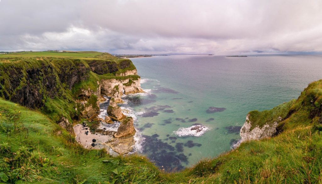 A scenic coastal landscape with rugged cliffs, a green grassy foreground, and calm blue-green sea waters under a cloudy sky, showcasing natural beauty and serenity.