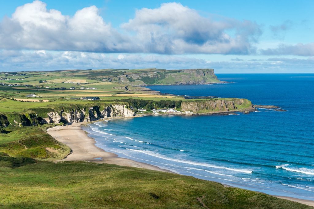 A panoramic view of a coastal landscape featuring a sandy beach, rugged cliffs, blue seas, green fields, and a partly cloudy sky above.