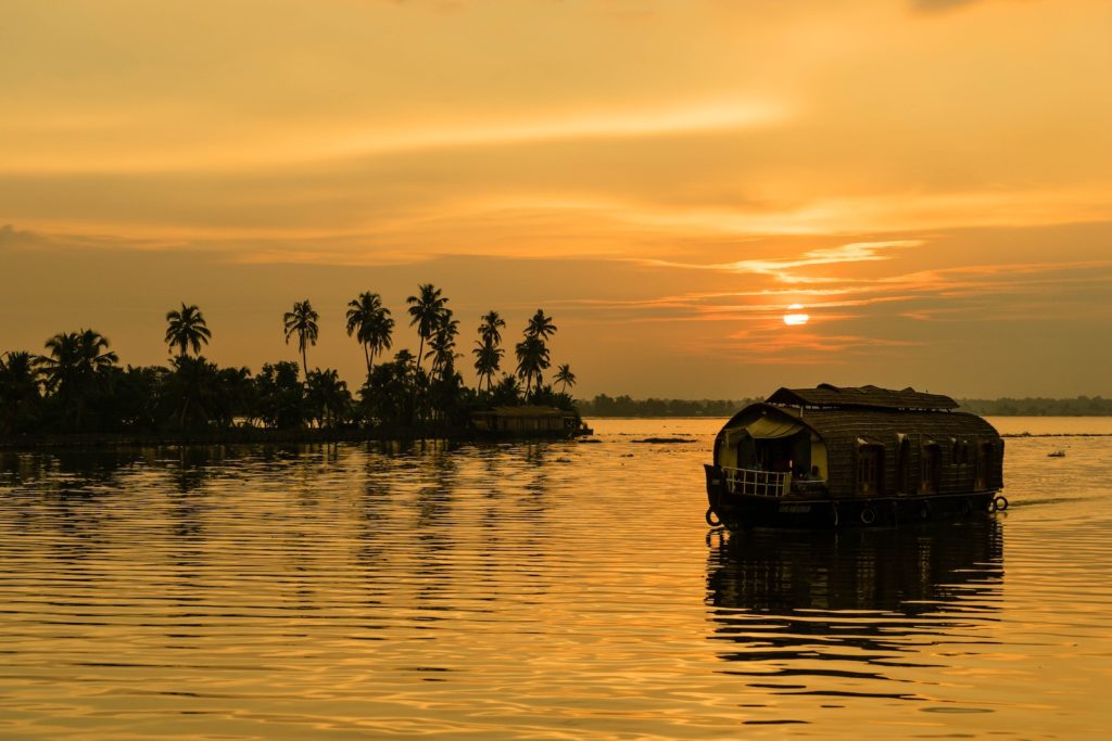 A houseboat floats on calm waters during sunset, with the sun casting a warm glow and silhouetting palm trees on a tranquil island landscape.