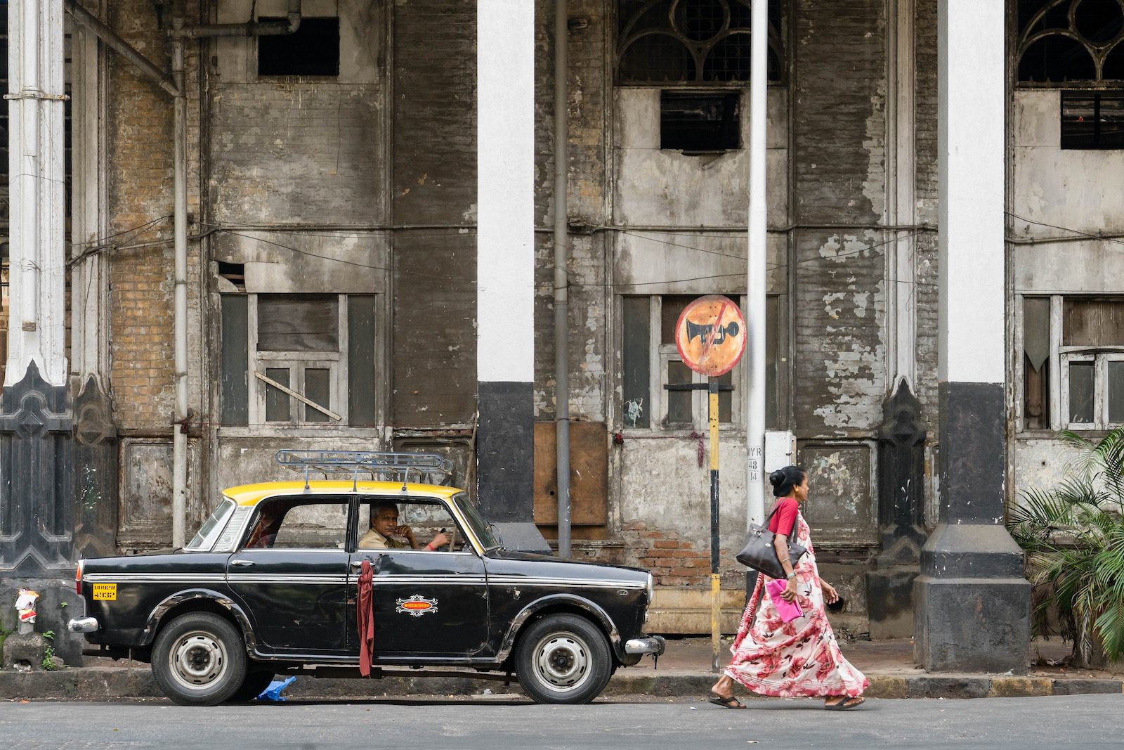 An old building with arched windows stands behind a parked taxi. A person walks past, wearing a patterned saree. A faded no parking sign is visible.