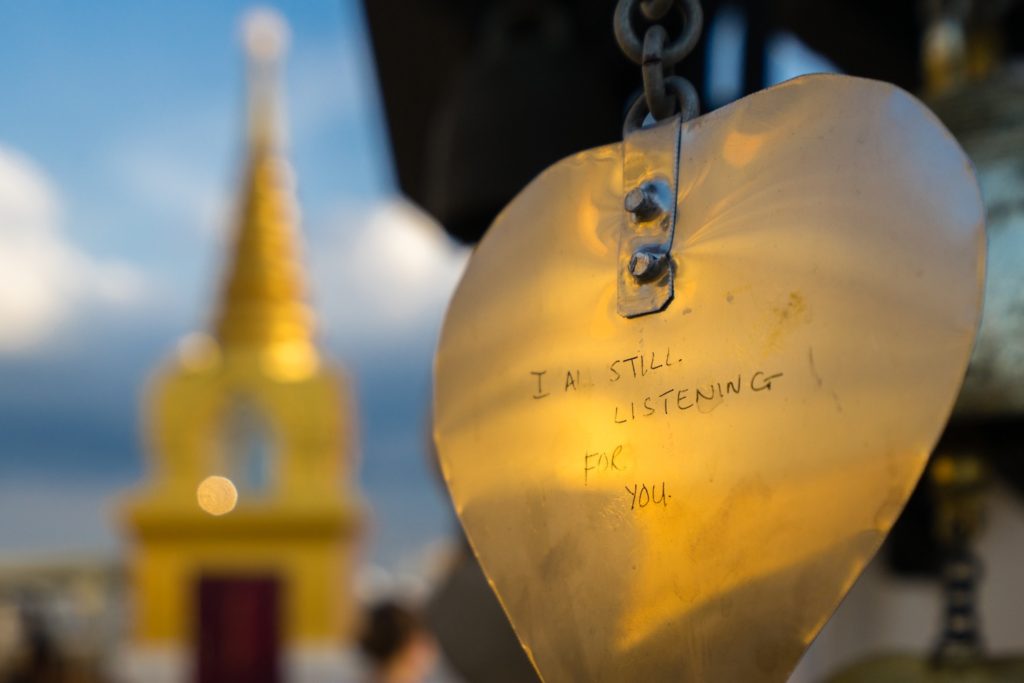 A golden heart-shaped object with the inscription "I AM STILL LISTENING FOR YOU" hangs by a chain, with blurred architectural structures in the background.