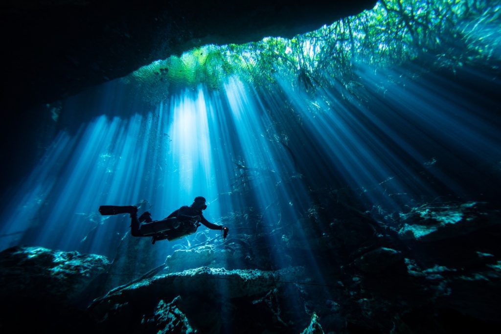 A person is scuba diving underwater in a cave, with light beams streaming through the water from an opening above, creating a serene, otherworldly scene.
