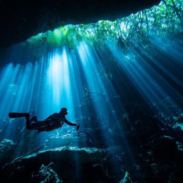 A person is scuba diving underwater in a cave, with light beams streaming through the water from an opening above, creating a serene, otherworldly scene.