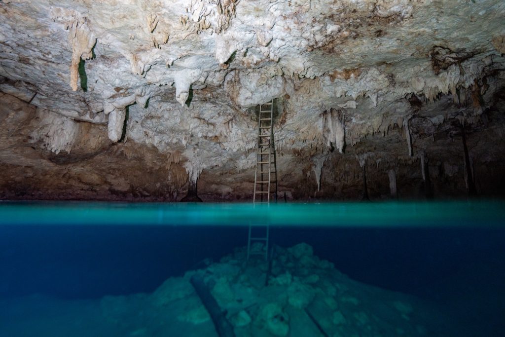 A wooden ladder extends from a rocky ledge into the clear blue water inside a cavern adorned with stalactites, suggesting a serene underground aquatic scene.