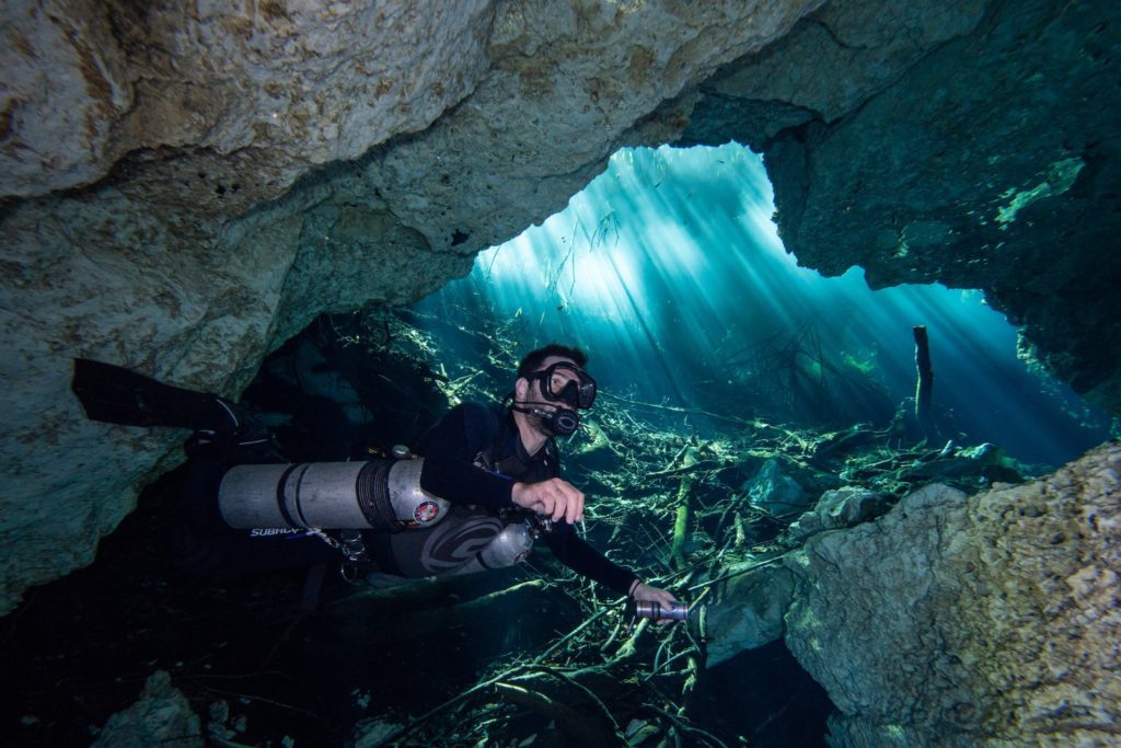 A person is cave diving, exploring an underwater cavern. Sunlight streams through an opening, illuminating the water and submerged rocks and plants.