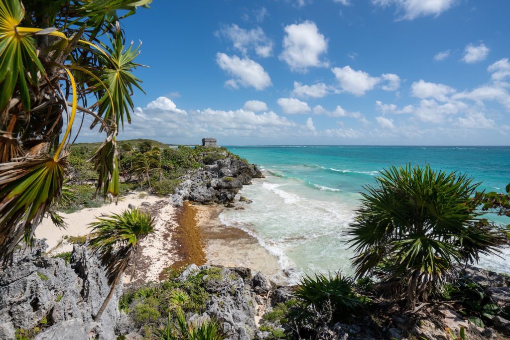 A tropical coastline with turquoise waters, white sandy beach, and rocky cliffs, framed by green foliage, under a blue sky with scattered clouds.