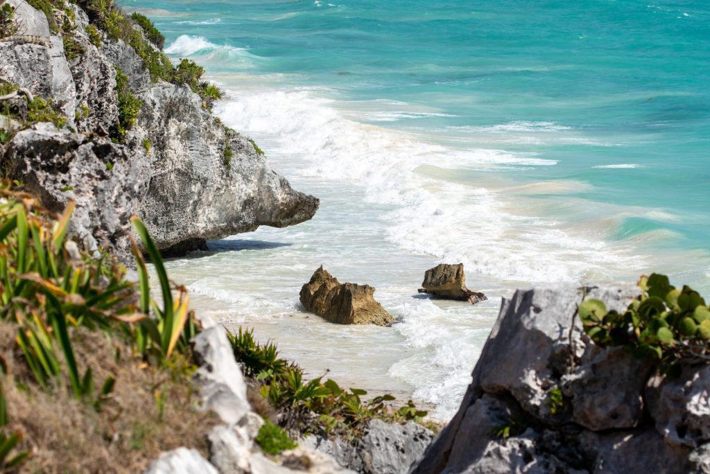 A tropical seascape with turquoise waters, white sandy beach, and rugged cliffs partially framed by green vegetation under a bright, sunny sky.