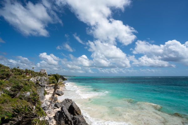 This image showcases a stunning coastal landscape with turquoise waters, a white sandy beach, and rocky cliffs under a vivid blue sky dotted with fluffy clouds.