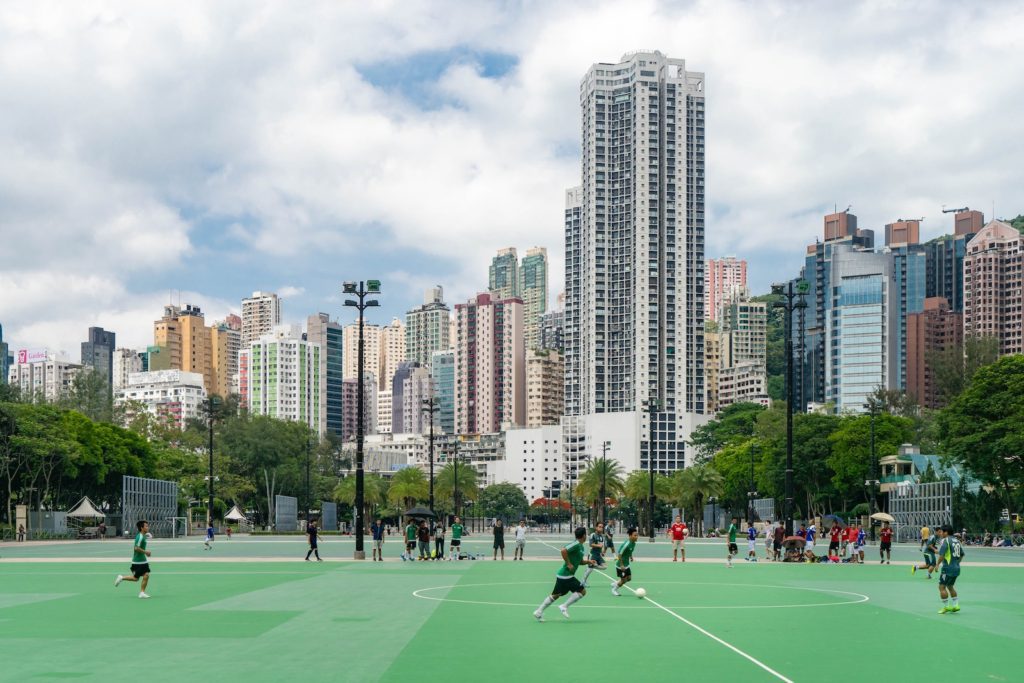 An outdoor sports field with several people playing a game, surrounded by tall city buildings under a partly cloudy sky.