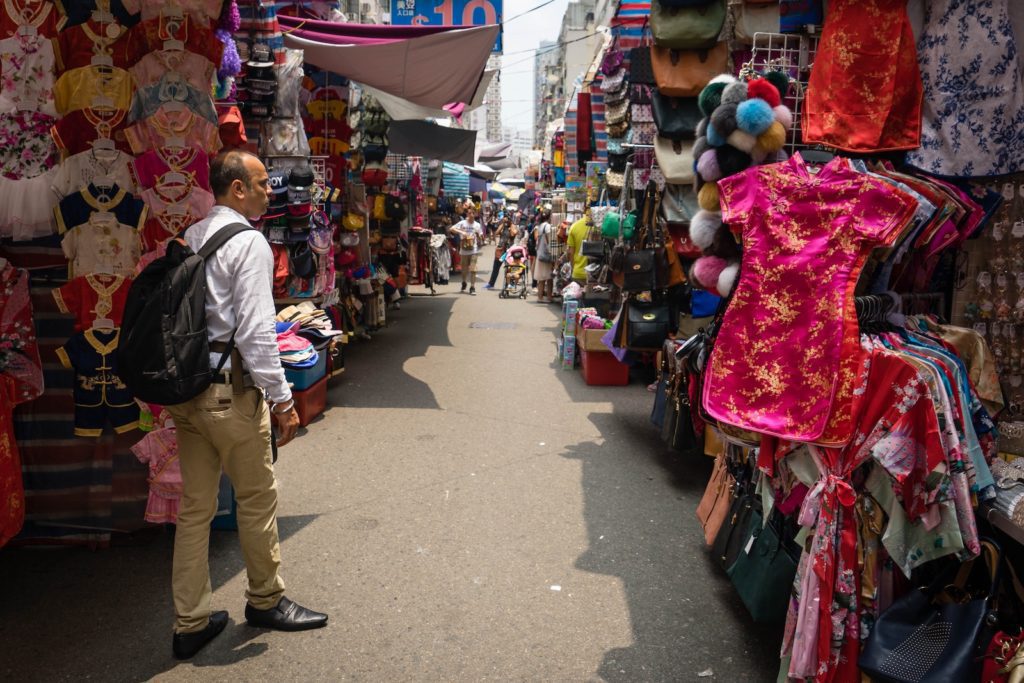 A person is standing in a bustling street market with clothes displayed on either side under bright sunlight, creating a lively shopping atmosphere.