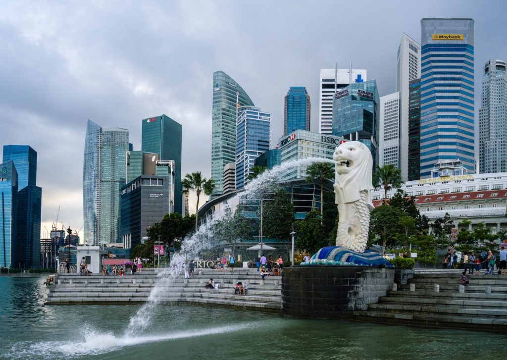 This image shows Singapore's iconic Merlion statue spouting water with the city's modern skyscraper-filled skyline in the background under a cloudy sky.