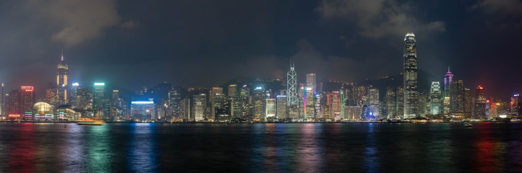 This image shows a panoramic night view of a vibrant city skyline with illuminated skyscrapers reflected in the water, presumably photographed from across a bay.