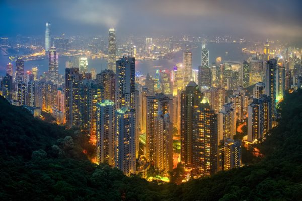 An evening view of a dense, brightly-lit urban skyline with skyscrapers, surrounded by dark hills with a misty atmosphere, showcasing a bustling metropolis.