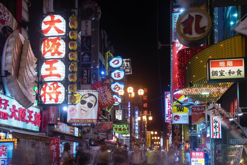 A bustling urban night scene with vibrant neon signs and billboards in Japanese script. Blurred figures suggest a crowd of people walking along the lively street.