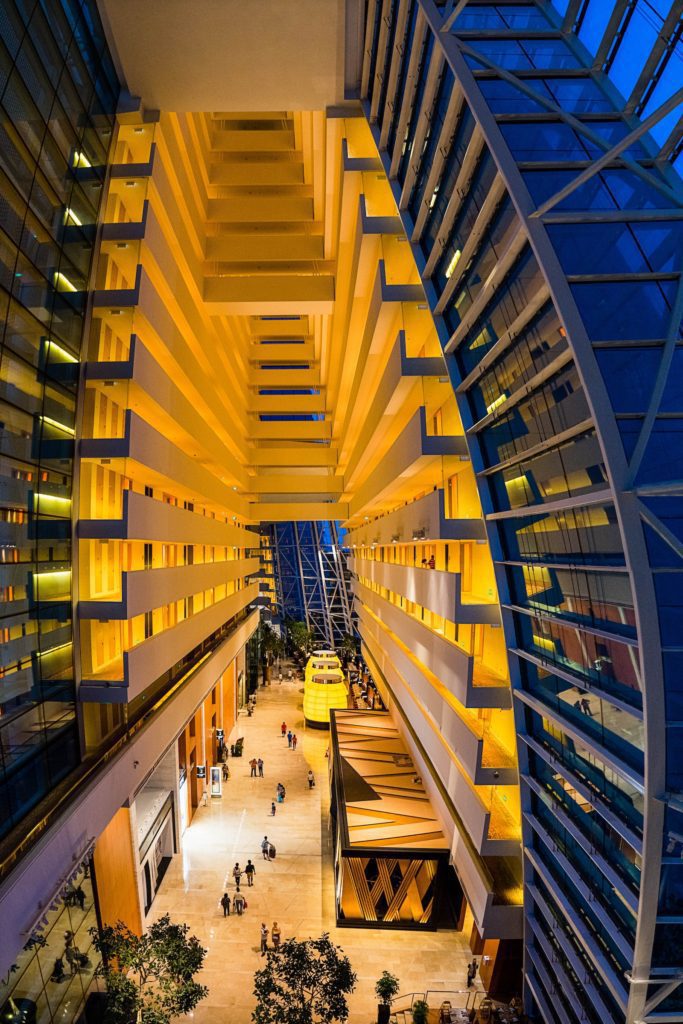 This image shows a towering modern interior atrium with warm yellow lighting, featuring balconies, glass elements, people walking, and a yellow tram.