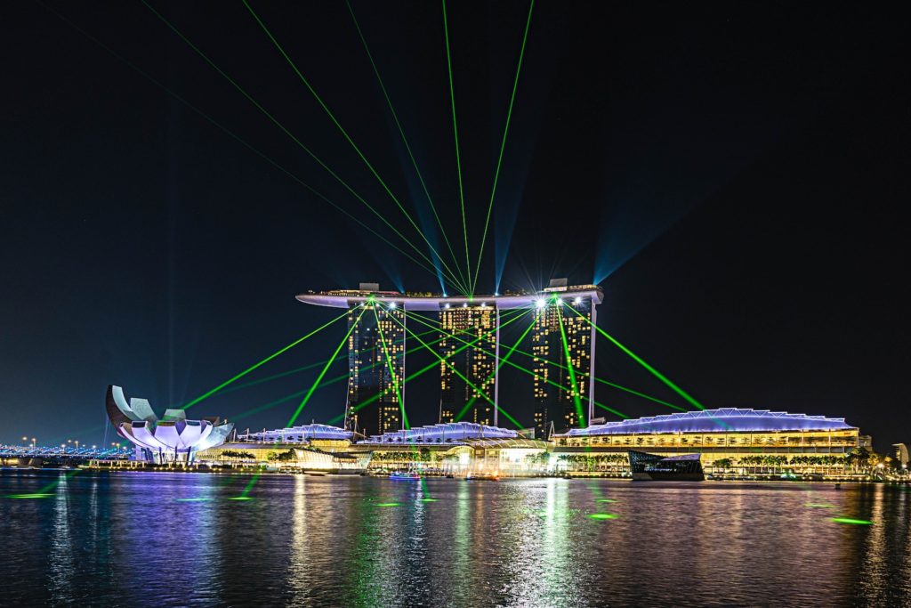The image shows the Marina Bay Sands hotel in Singapore at night with a light show featuring green lasers emanating from its rooftop.