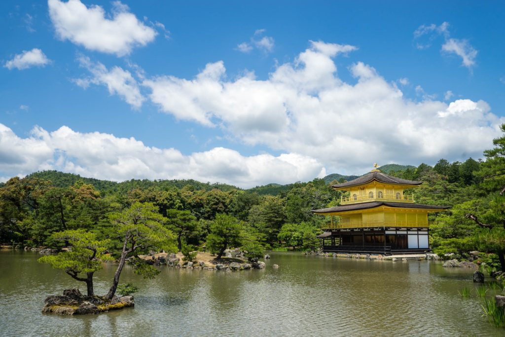 Golden pavilion reflects in a tranquil pond surrounded by lush greenery under a partly cloudy sky. Traditional Japanese architecture amidst a serene landscape.