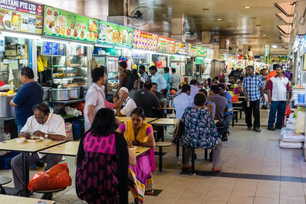 This image shows a busy indoor food court with various stalls. People are dining at tables, walking, and ordering food. The atmosphere seems lively.