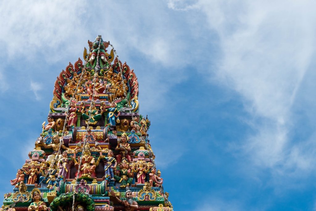 The image shows a colorful, ornate gopuram, a traditional tower at the entrance of a Hindu temple, set against a bright blue sky with light clouds.