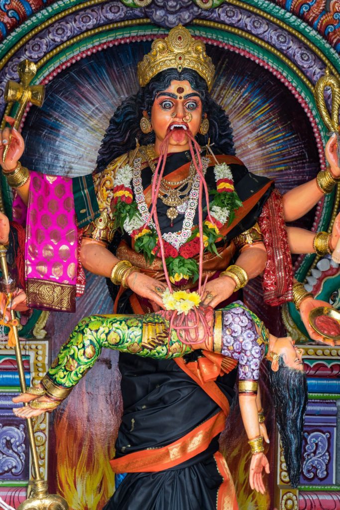 This is a colorful statue of the Hindu goddess Kali, with multiple arms, standing on a figure, against a vibrant circular backdrop. She wears traditional attire and jewelry.
