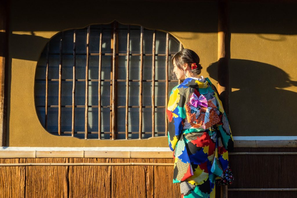 A person stands beside a traditional Japanese window, back facing the camera, wearing a colorful, patchwork kimono, in the warm light of sunset.