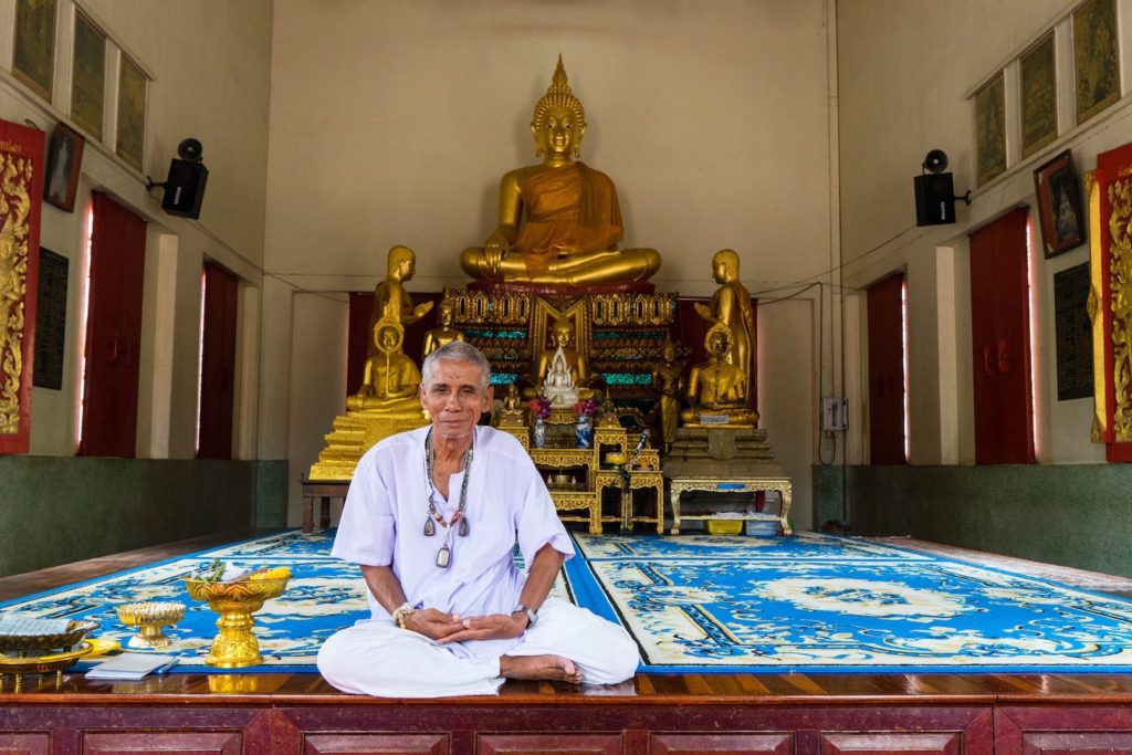 A person is sitting cross-legged on a blue mat inside a temple, in front of a large golden Buddha statue, with a serene expression on their face.