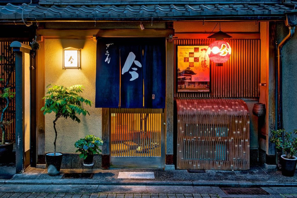 A cozy Japanese street at dusk featuring a traditional restaurant facade with a noren curtain, illuminated lanterns, and bamboo blinds, radiating a warm, welcoming atmosphere.
