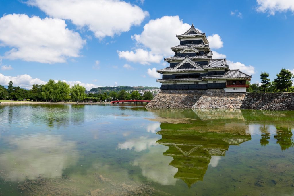 A multi-tiered traditional Japanese castle with a stone foundation reflects onto the calm water before it, under a blue sky with fluffy clouds.