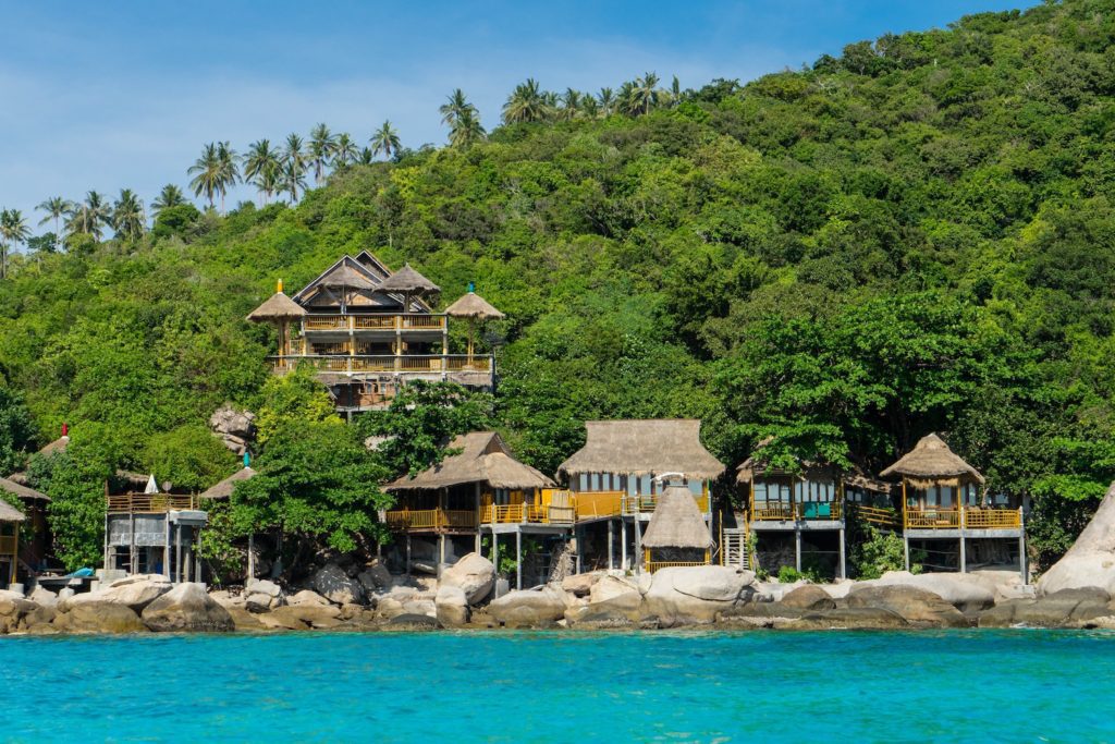 Tropical resort nestled on a hillside with traditional thatched-roof buildings overlooking a serene blue sea, surrounded by lush greenery under a clear sky.