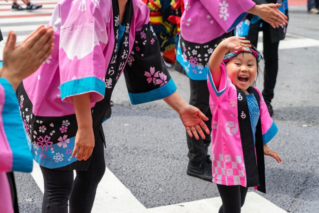 A joyful child in a pink hanbok is smiling and reaching out during a procession with adults in similar attire, possibly a cultural or festival event.