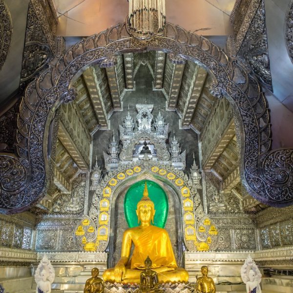 This image portrays a golden Buddha statue seated inside an ornate temple with intricate silver artwork, surrounded by smaller figurines and elaborate decorations.