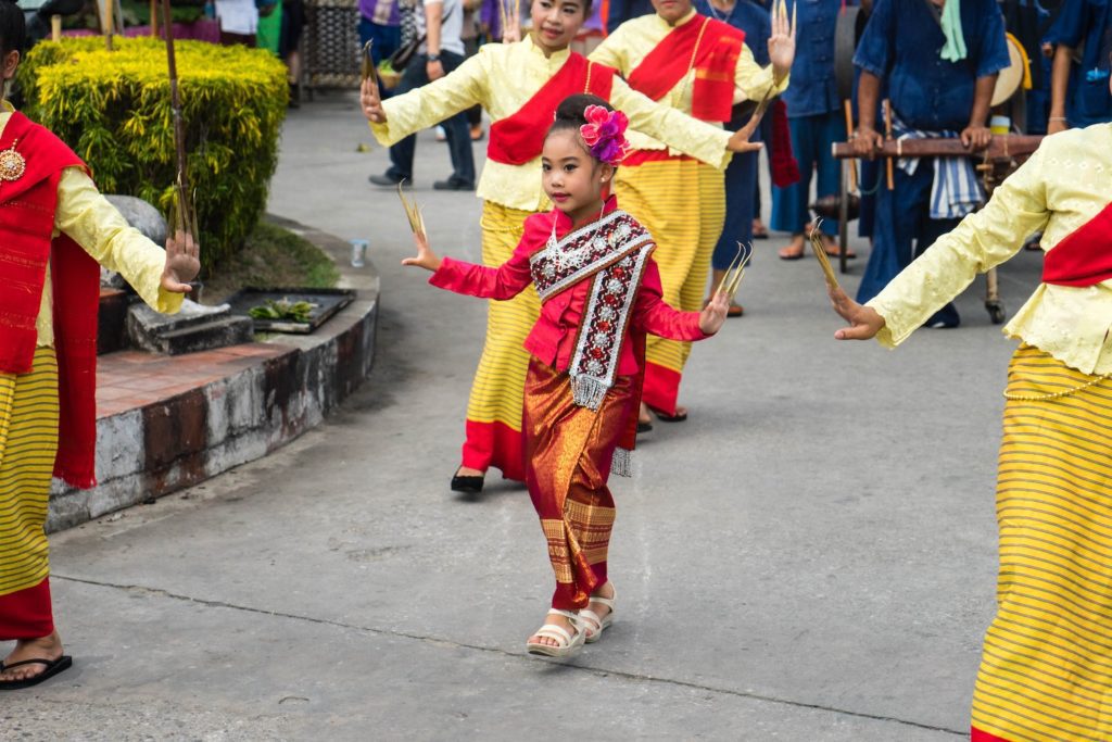 A young child dressed in traditional colorful attire performs a dance on a street, surrounded by people in similar costumes, likely at a cultural festival.