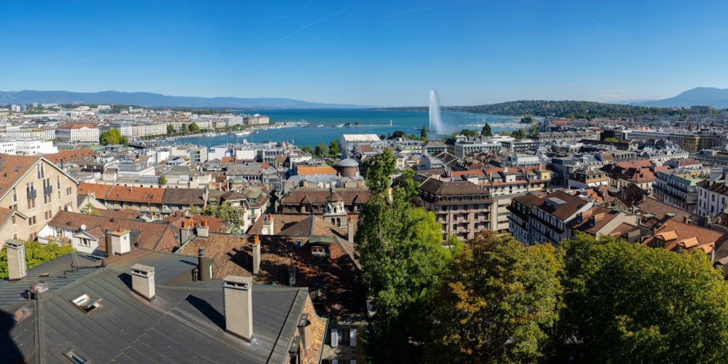 This panoramic image features a cityscape with traditional European architecture, a large water fountain jet in a lake, and hills in the background under a clear sky.