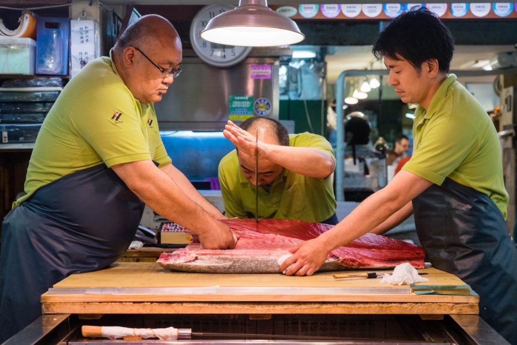 In a bustling market, two people in green uniforms meticulously prepare a large fish on a wooden table, under bright lights, suggesting expertise in seafood handling.