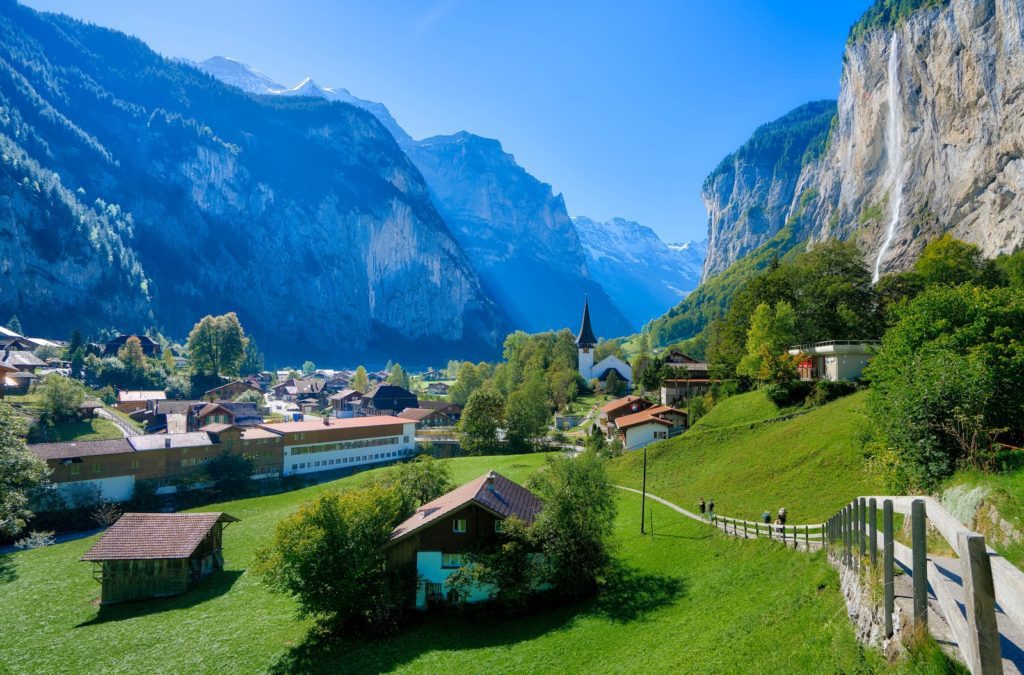 A picturesque village nestled in a valley with lush green fields, surrounded by towering cliffs and alpine mountains under a clear blue sky.