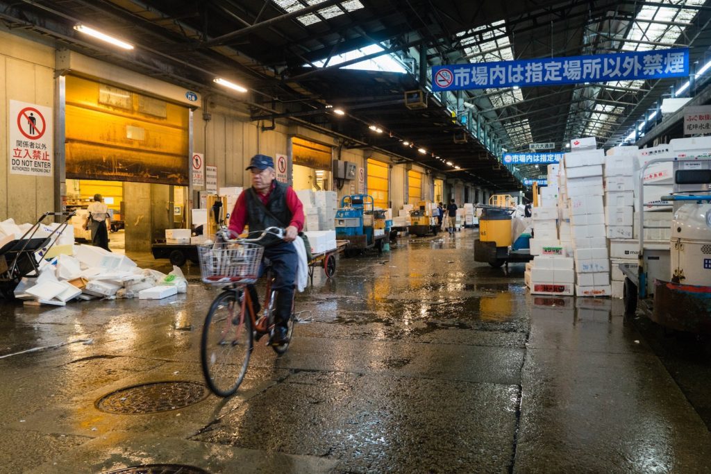 A person cycles through a wet indoor market with carts and boxes. Signs and infrastructure suggest it is likely in an East Asian location.