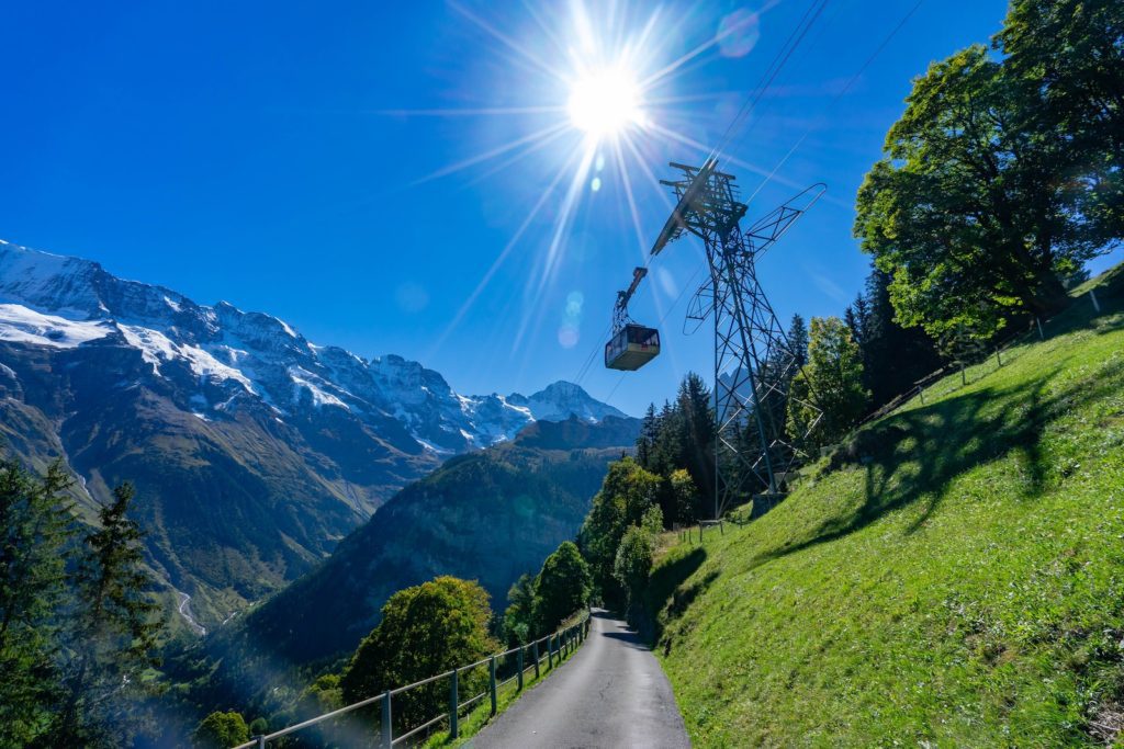 A cable car descends over a lush green hillside with a clear path in the foreground and snow-covered mountains under a bright sun in the background.