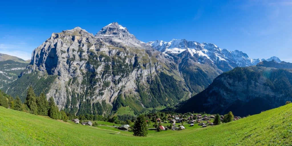 A panoramic view of a lush green valley with a small village, framed by majestic snow-capped mountains and a clear blue sky.