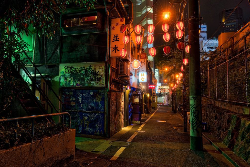 An atmospheric alley at night, illuminated by red lanterns and neon signs, with graffiti on the left wall and a cityscape in the background.