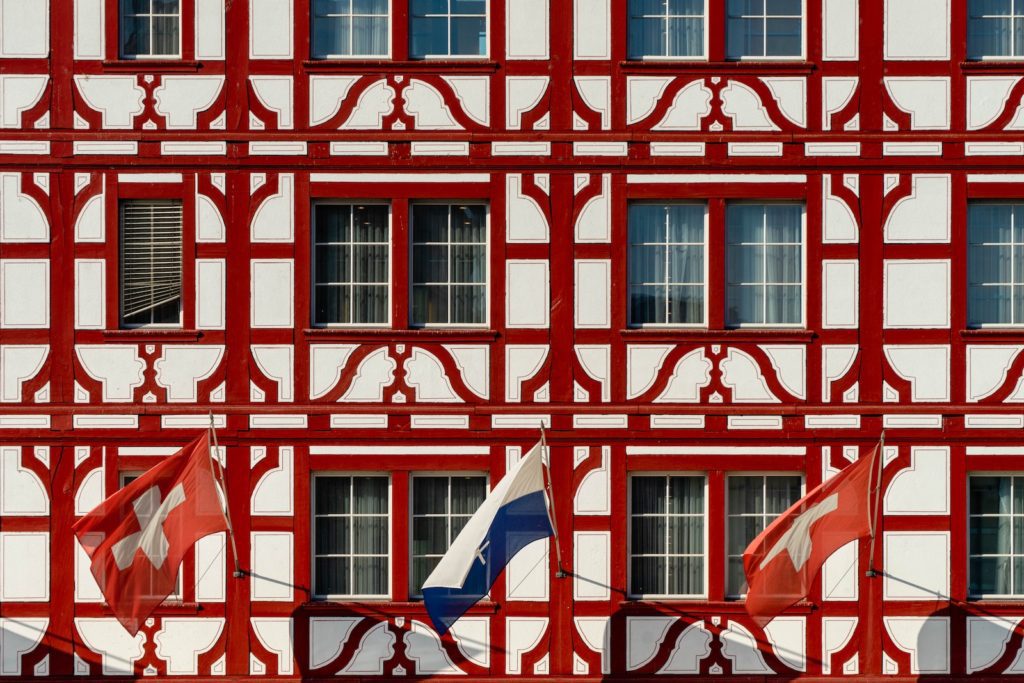 The image shows a section of a red and white half-timbered building façade with windows and two flags, one likely representing Switzerland, fluttering in the breeze.