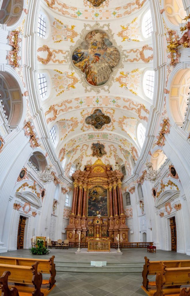 Baroque style church interior featuring ornate gold altar, frescoed ceilings with elaborate stucco, white walls, arched aisles, and wooden pews in foreground.