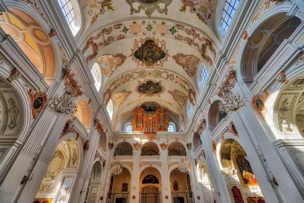 This is an ornate baroque church interior, featuring elaborate ceiling frescoes, architectural columns, arched windows, and an intricate pipe organ visible at the back.